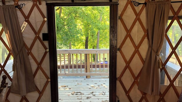 Una carpa con piso de madera, vista a árboles y una cerca en el exterior, y una felpudo con un diseño.