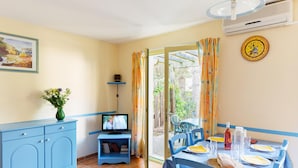 A room with a dining table set for two, a television on a stand, a vase with flowers, and a clock on the wall. - Charming Countryside Maisonette (Les Mathes)
