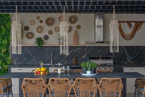 A modern kitchen with a marble backsplash, hanging macramé lights, and wicker chairs around a central island with fruit and a wine bottle. - Azur BlueSky luxury villa , great views near the beach (Bali)