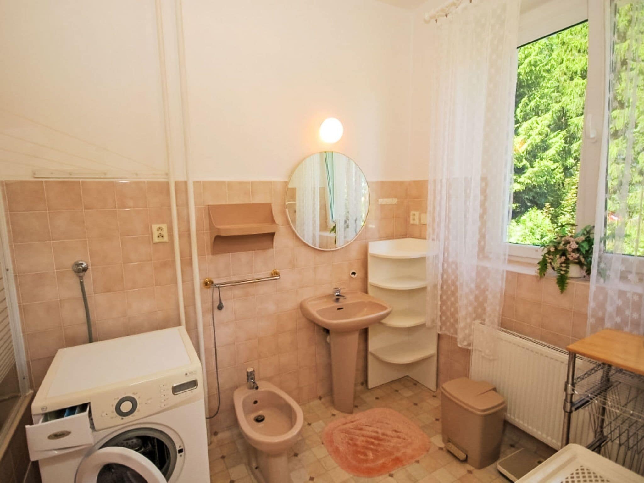 A bathroom with a washing machine, bidet, sink, and a window with a view of greenery.