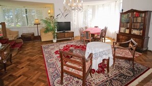 Una sala de estar con televisor, candelabro, mesa de comedor con sillas, estantería y un amplio ventanal con cortinas.