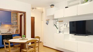 A modern kitchen with blue cabinets, a dining table with chairs, a TV on a white stand, and a small potted plant. - Casa Elena by Interhome (Bellano)