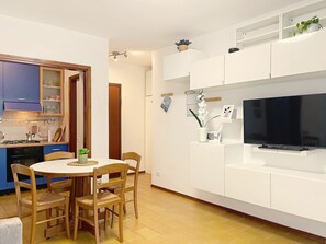A modern kitchen with blue cabinets, a dining table with chairs, a TV on a white stand, and a small potted plant. - Casa Elena by Interhome (Bellano)