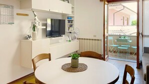 A modern living room with a TV, white furniture, a dining table with chairs, a small plant, and a view of the outside. - Casa Elena by Interhome (Bellano)