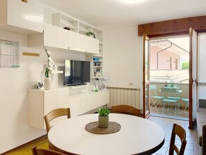A modern living room with a TV, white furniture, a dining table with chairs, a small plant, and a view of the outside. - Casa Elena by Interhome (Bellano)
