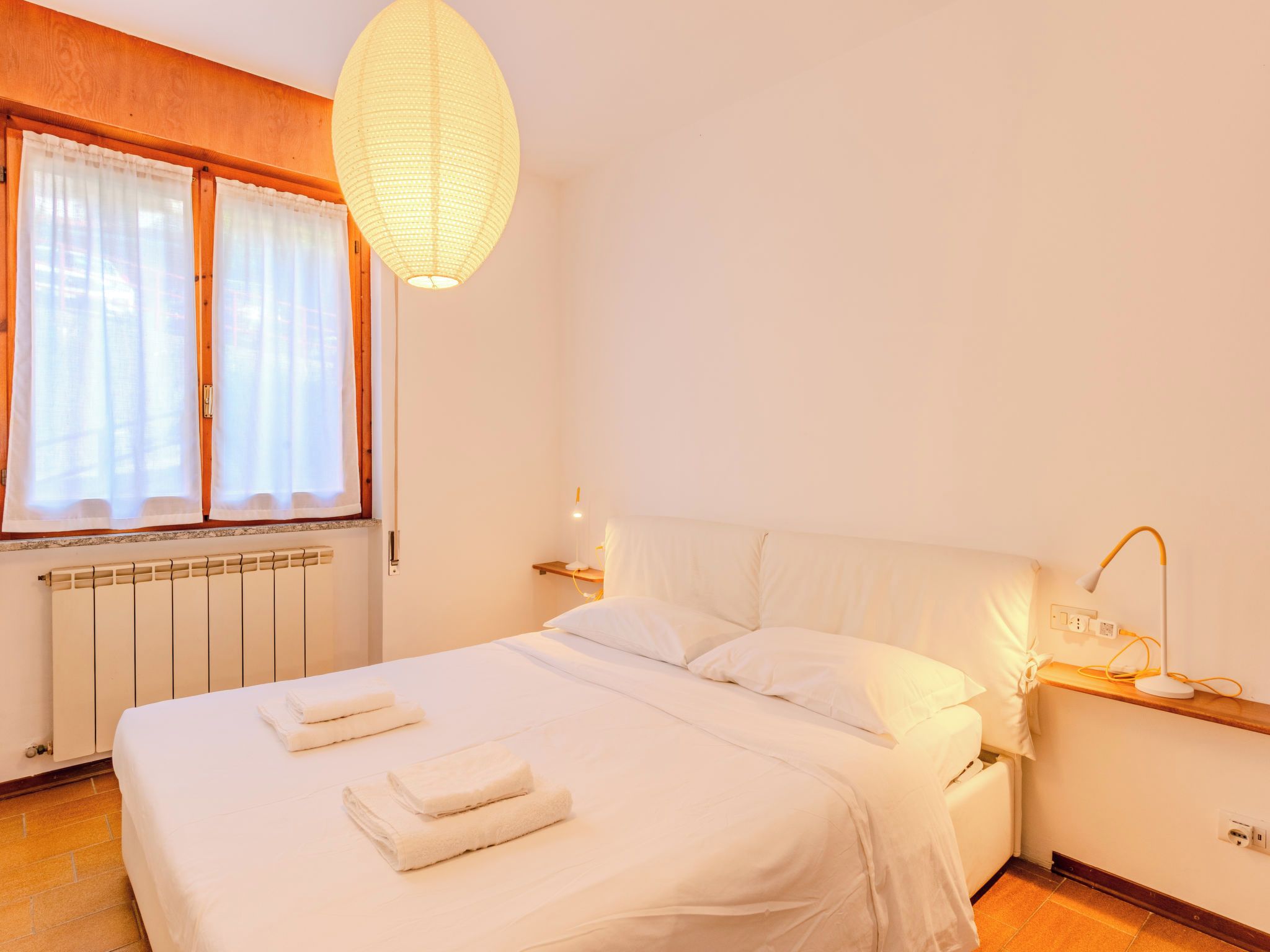 A hotel room with a bed, a bedside table with a lamp, a window with curtains, and a radiator.