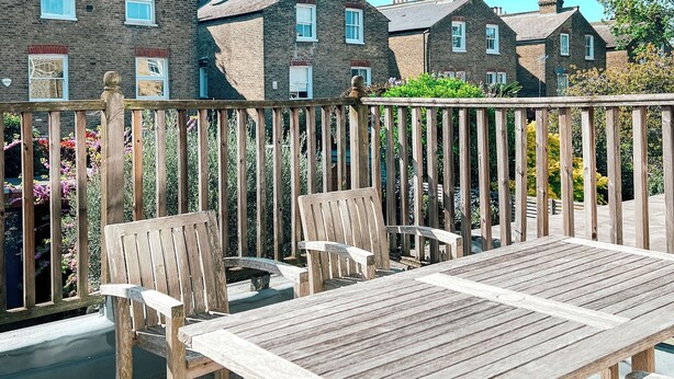 A wooden deck with a table and chairs, a wooden fence, and brick houses in the background. - Sugden Road (London)