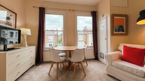 A room with a dining table, chairs, a TV, a lamp, a sofa with a red pillow, and a window with curtains. - Sugden Road (London)
