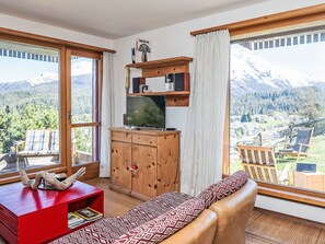 Ein Wohnzimmer mit einer Couch, einem Fernseher, einer Holzkommode und Blick auf Berge.