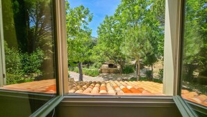 Näkymä ikkunasta terassialueelle, jossa on kivinen takka ja jota ympäröivät puut ja vihreys.