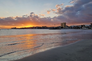 Ein Strand mit Sonnenuntergang, ruhigem Wasser und Gebäuden im Hintergrund.