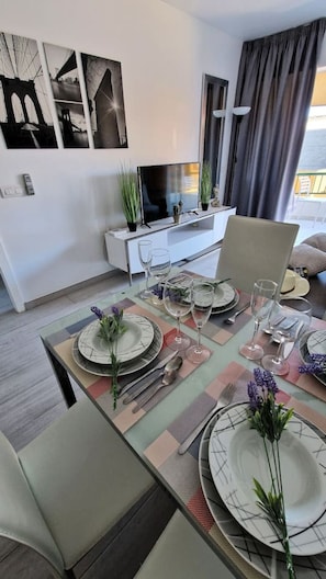 Ein Essbereich mit einem Tisch für vier Personen, einem Fernseher an einer Wandhalterung und einer Couch im Hintergrund.
