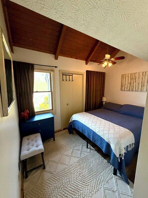 A bedroom with a bed, a desk, a chair, a ceiling fan, and a window.