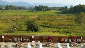 A swimming pool with lounge chairs and umbrellas, surrounded by a wooden fence and a grassy field with trees and hills in the background.
