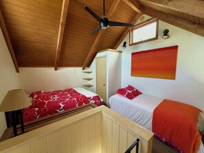 Ein Schlafzimmer mit schrägverdeckter Decke, einem Deckenventilator, einem Bett mit rotem floralem Überwurf, einem Nachttisch mit Lampe, einem Fenster und einem Wandbild.