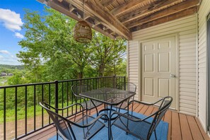 Eine überdachte Terrasse im Freien mit Tisch und Stühlen, einer Tür und Blick auf Bäume sowie ein Haus in der Ferne.
