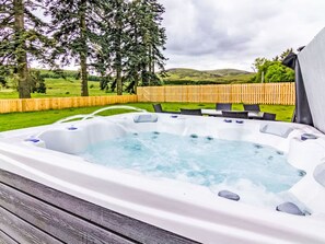 Un bain à remous doté d’une terrasse en bois, entouré d’une clôture en bois et d’arbres, avec un champ herbeux et des collines en arrière-plan.