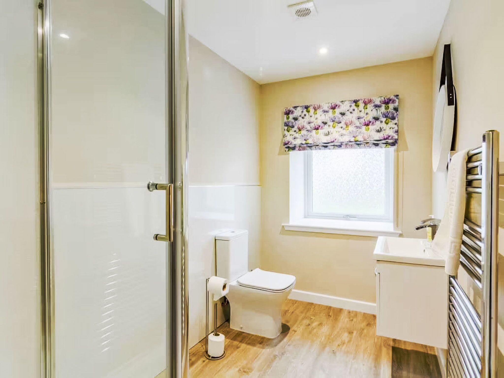 Een badkamer met een toilet, een douche, een raam met bloemen gordijn en een handdoekhouder.