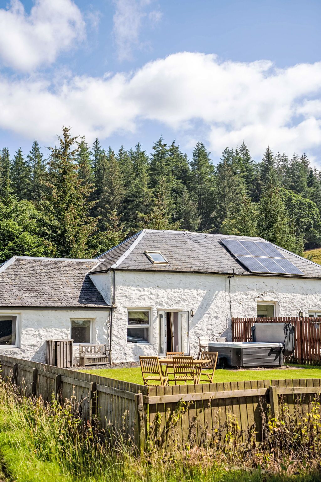 A white stone building with a sloped roof, solar panels, and a fenced yard with a table and chairs.