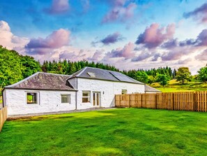 Une maison blanche avec un toit en pente, une cour clôturée et, en arrière-plan, des arbres et un ciel nuageux.