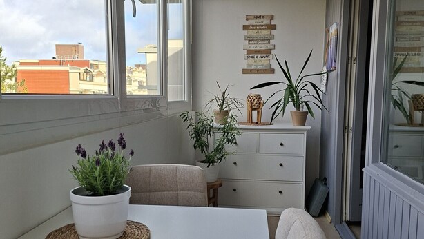 Ein Balkon mit weißem Tisch, gewobenen Unterlagen, Topfpflanzen, einer weißen Kommode und einer Wand mit inspirierenden Zitaten.