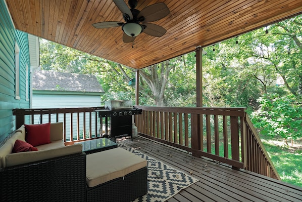 A covered outdoor deck with a ceiling fan, a grill, and a seating area with cushions.