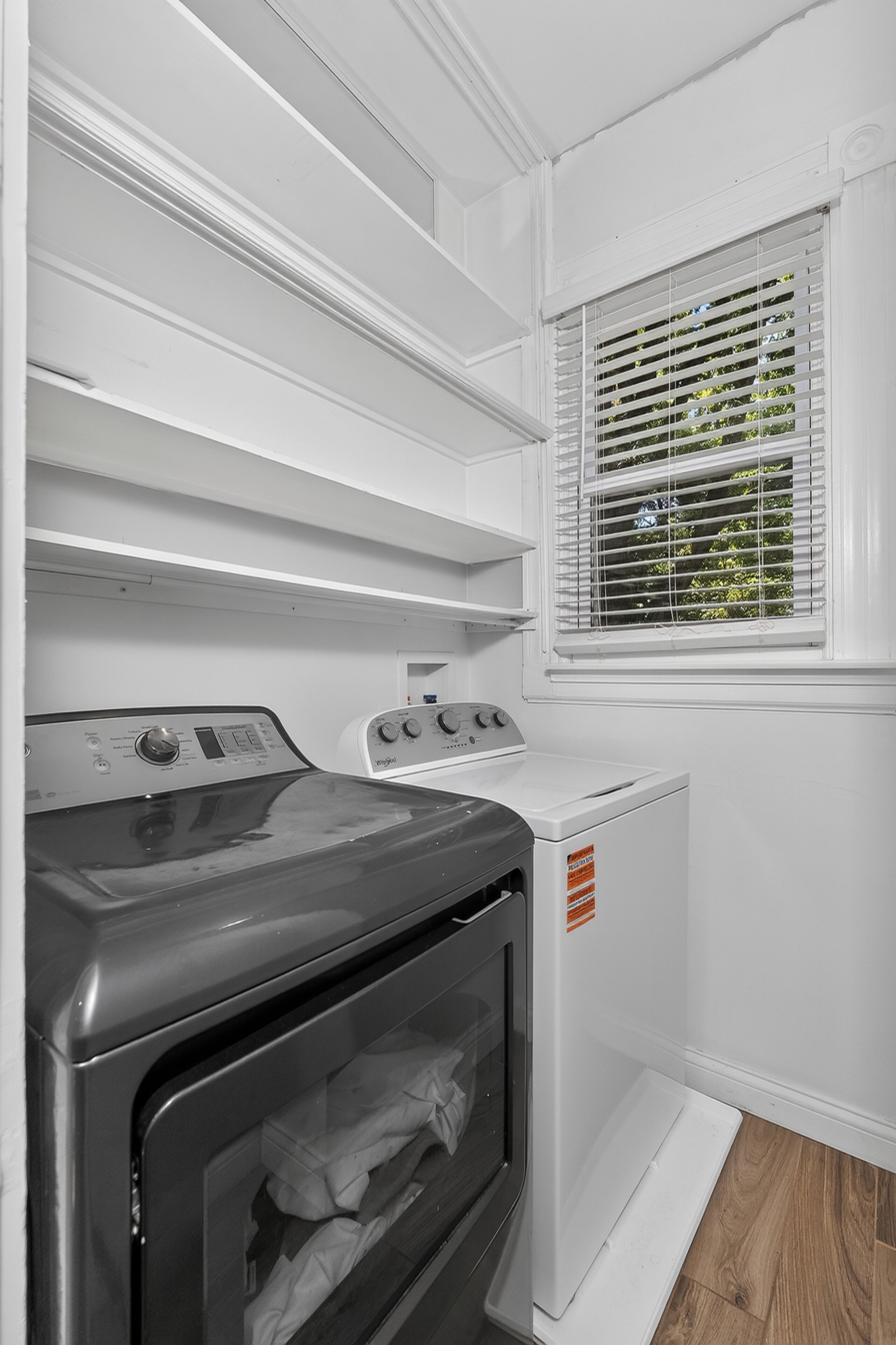 A laundry room with a front-loading washer and dryer, a window with blinds, and built-in shelving above the appliances.