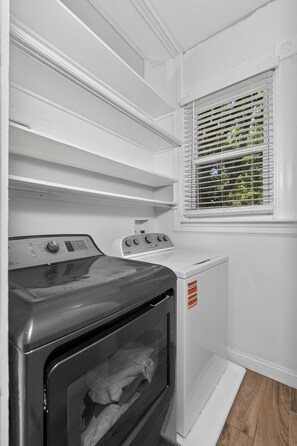 A laundry room with a front-loading washer and dryer, a window with blinds, and built-in shelving above the appliances.