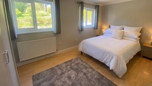 Een slaapkamer met een groot bed, twee ramen met gordijnen, een radiator en een grijs vloerkleed.