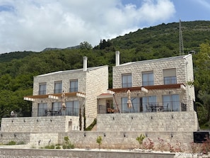 A two-story stone building with balconies, outdoor seating, and a view of a green hillside.