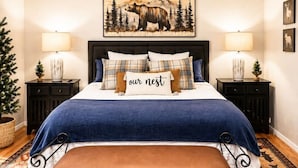 A bedroom with a bed, a bench, a ceiling fan, two bedside tables with lamps, a wall-mounted painting, and a patterned rug. - Alpine Escape in Ruidoso NM (Ruidoso)