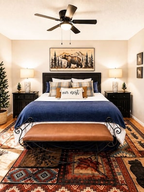 A bedroom with a bed, a bench, a ceiling fan, two bedside tables with lamps, a wall-mounted painting, and a patterned rug. - Alpine Escape in Ruidoso NM (Ruidoso)