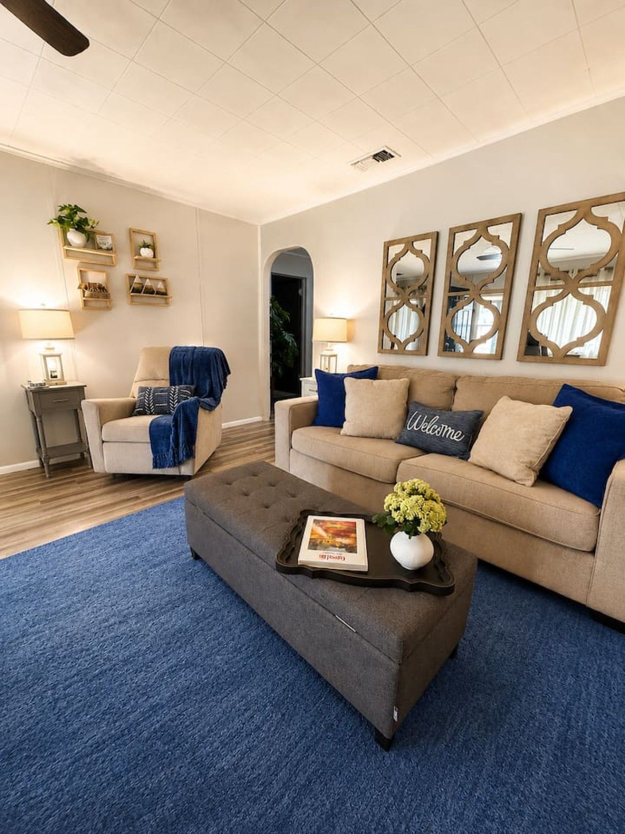 Un salon comprenant un canapé, un fauteuil et un pouf, une table basse avec un magazine et des fleurs, ainsi que des miroirs décoratifs accrochés au mur.