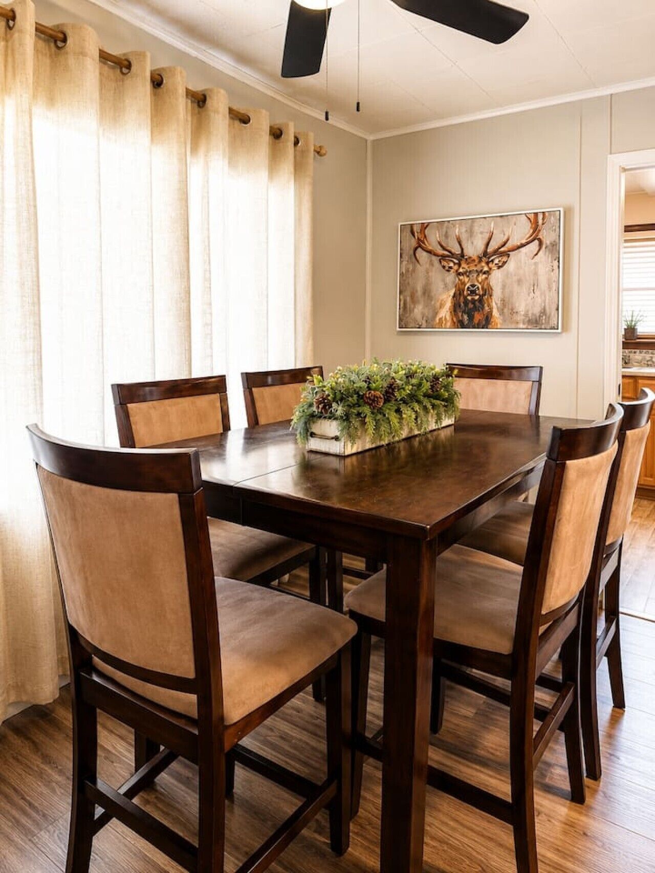 A dining room with a wooden table, six chairs, a decorative plant centerpiece, and a deer painting on the wall.