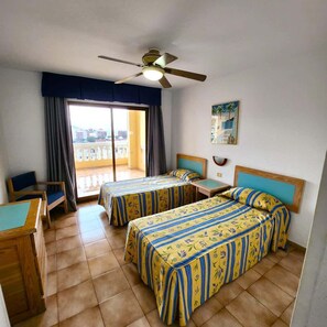 A hotel room with two beds, a desk, a chair, a ceiling fan, and a balcony with curtains. - Castle Harbour 40 - One Bed with Sea View (Los Cristianos)
