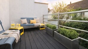 A balcony with a blue sofa, yellow cushions, a small table with a pitcher and glasses, and planters with greenery. - The Cornstore -  a house that sleeps 6 guests  in 3 bedrooms (Southwold)