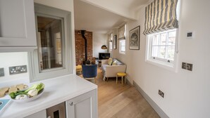 A modern kitchen with a white countertop, a bowl of vegetables, a brick wall, a sofa, and a window with striped curtains. - The Cornstore -  a house that sleeps 6 guests  in 3 bedrooms (Southwold)