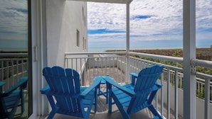 Een balkon met twee blauwe Adirondack-stoelen en een tafel, met uitzicht op een strand en de oceaan.