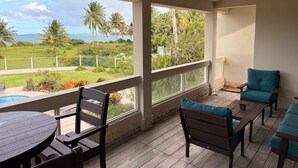 A covered outdoor area with a pool, wooden furniture, and a view of a grassy area with palm trees and a body of water. - Beachfront, Luxury Oasis with Private Resort Pool (Palmer)