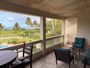 A covered outdoor area with a pool, wooden furniture, and a view of a grassy area with palm trees and a body of water. - Beachfront, Luxury Oasis with Private Resort Pool (Palmer)