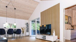 Ett modernt vardagsrum med ett träpanelat tak, en platt-TV, en matplats med stolar och ett kök med träpanel.