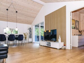 Ein modernes Wohnzimmer mit Holzboden, einem Flachbildfernseher, einem Essbereich mit Stühlen und einer Küche mit Holzvertäfelung.