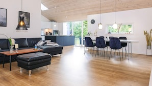 Ett modernt vardagsrum med ett träpanelat tak, en matplats med stolar, en soffa och ett kök i bakgrunden.