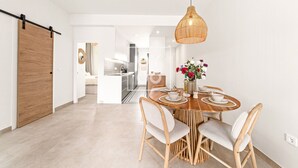 A modern kitchen with white cabinets, a wooden door, and a dining area with a wooden table and chairs. - Marina Garden Swim-Up Suite, direct pool access, colourful boho chic resort retreat - by OYOOSTAYS (Pilar de la Horadada)