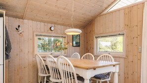 A wooden-paneled room with a dining table and chairs, two windows with views of greenery, a clock on the wall, and a hanging light fixture. - 4 Star Holiday Home in Lokken (Lokken)