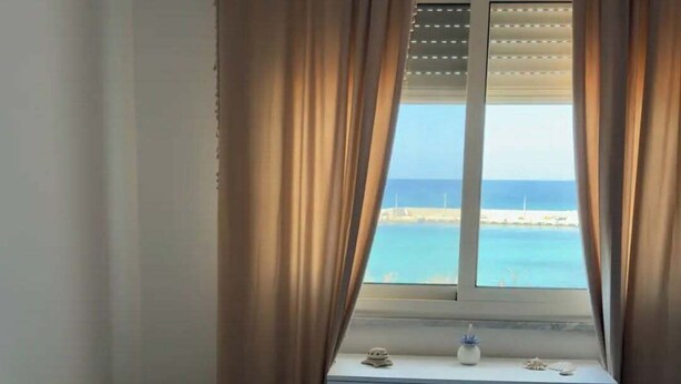 A bedroom with a bed, a window with curtains, a small table with decorative items, and a view of the sea. - Cozy 1-bedroom apartment in front of the sea (Palermo)