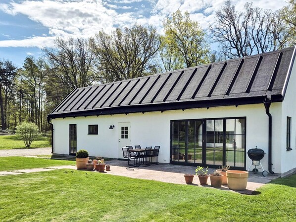 Un bâtiment blanc avec un toit noir, de grandes fenêtres et un patio aménagé avec du mobilier d’extérieur.
