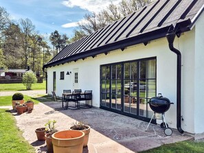 Un bâtiment blanc avec un toit noir, de grandes fenêtres en verre, des places assises à l’extérieur et un barbecue.