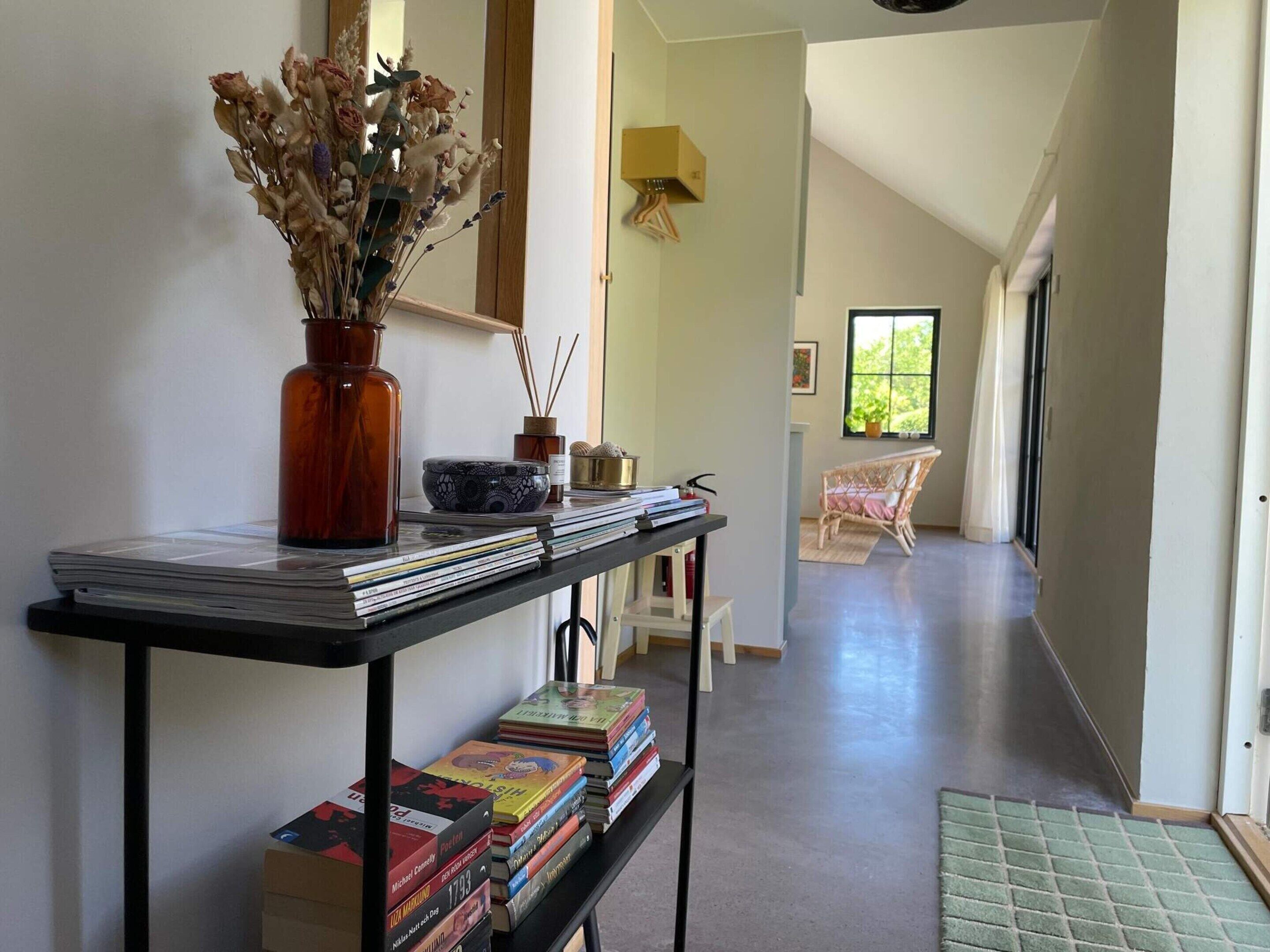 Un pasillo con una mesa que exhibe libros y un jarrón con flores secas, que conduce a una habitación con una silla de mimbre y un ventanal.