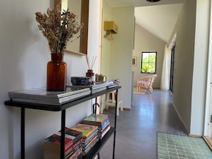 A hallway with a table displaying books and a vase of dried flowers, leading to a room with a wicker chair and a window. - 5 Star Holiday Home in Farjestaden (Farjestaden)